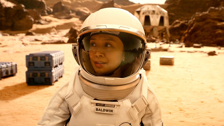 Person wearing a space suit and walking through a dry beige landscape